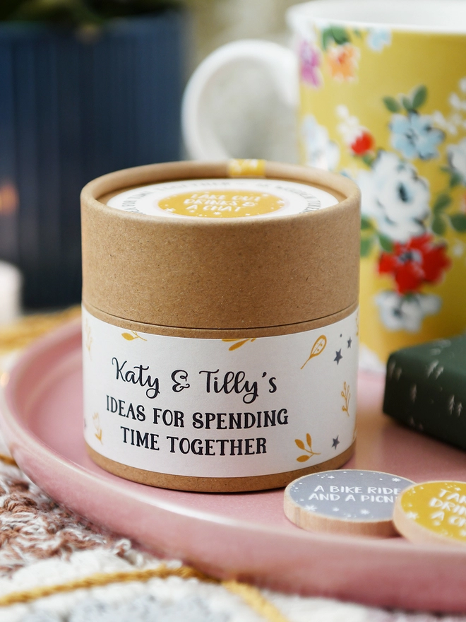 A cardboard jar with a floral label on the side that reads Ideas for spending time together stands on a pink plate.