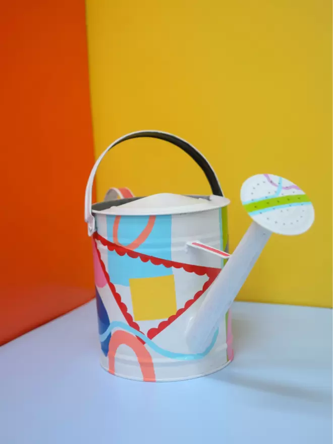 Hand painted watering can by Julie-Anne Pugh. Base colour is white with bring coloured shapes weaving across the body of the can on a yellow and orange background. Photographed from the front.
