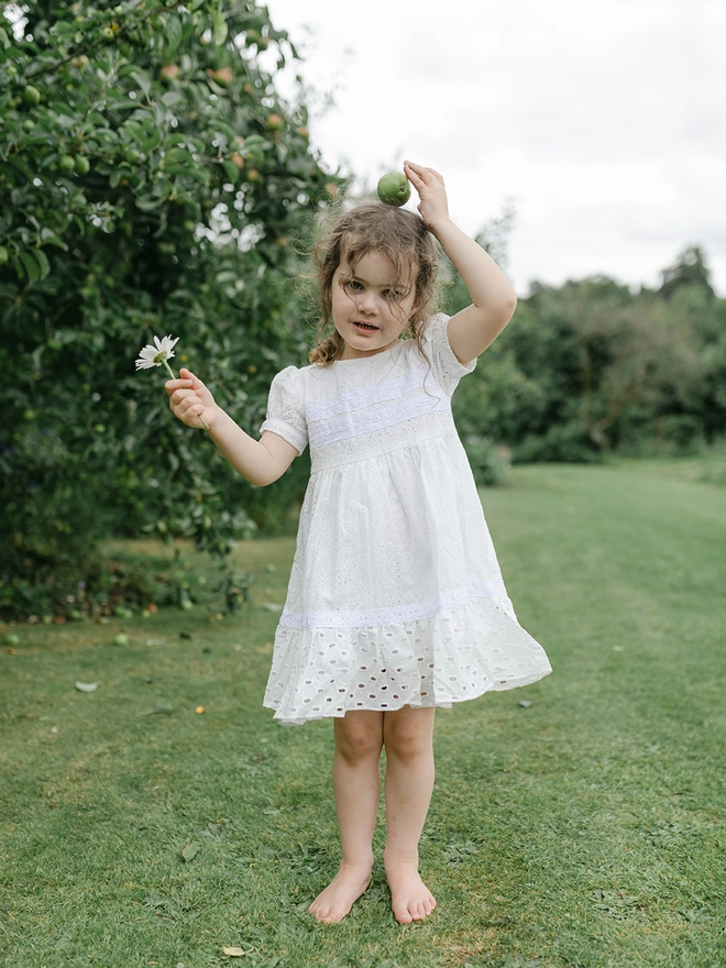 A little girl in a dress of contrasting white broderie anglaise fabrics stands beside a tree with an apple on her head