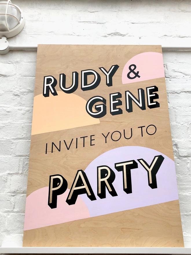 Handpainted invite to party sign with gold leaf letters outlined in black, with pastel coloured semicircles in the background, against a white brick wall. 