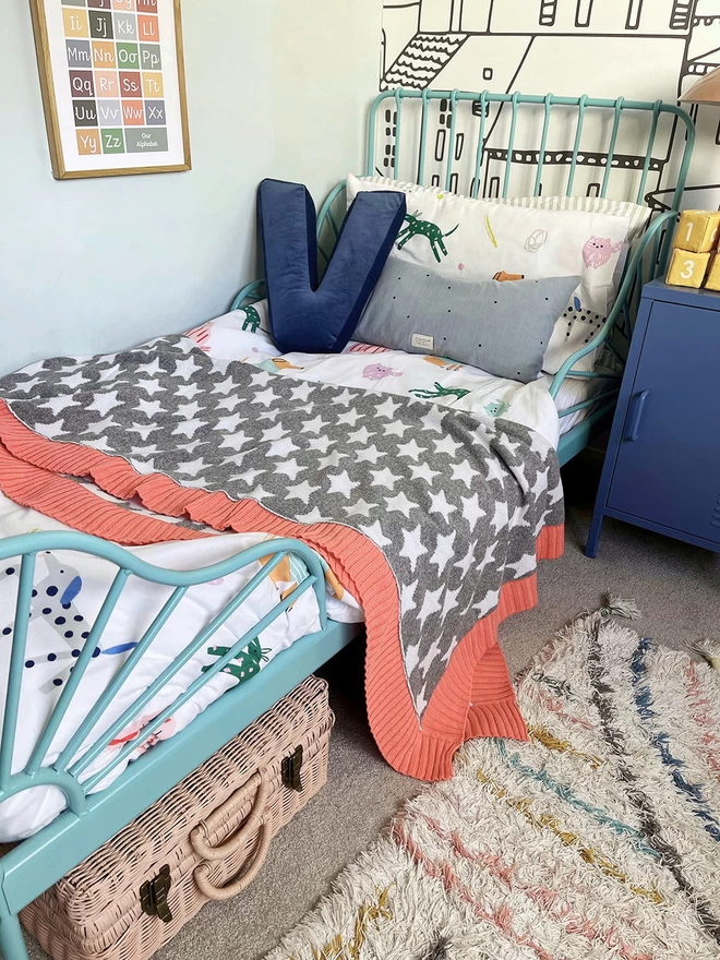 A view of a warm and vibrant childrens bedroom in muted rainbow colours showing a single bed with a grey and white star blanket with coral pink trim folded in half on the bed.