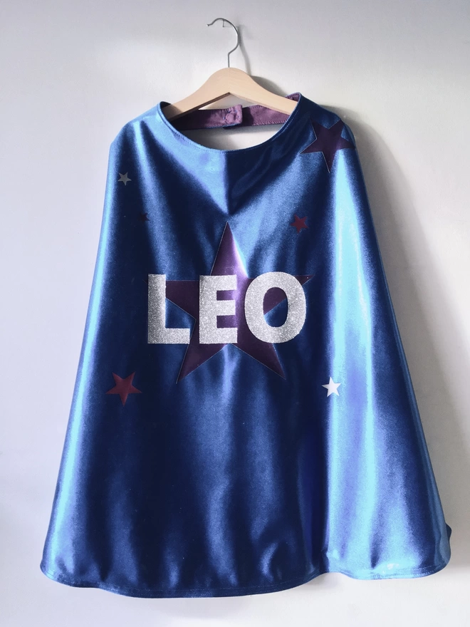 Sapphire Blue superhero cape with large purple star design and a name in silver glitter, hanging on a wall