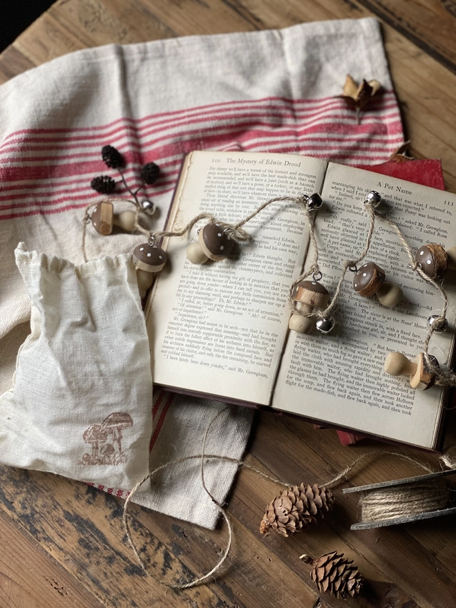 A string of Hand Painted Wooden Toadstool Bell Garlands trailing across an opened book atop a red and white striped cloth, on display with a small white pouch and some decorative pine cones