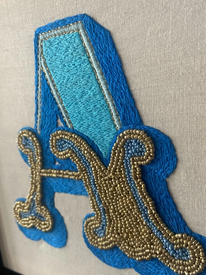 A close up of an embroidered letter A in shades of blue with gold and blue seed beads