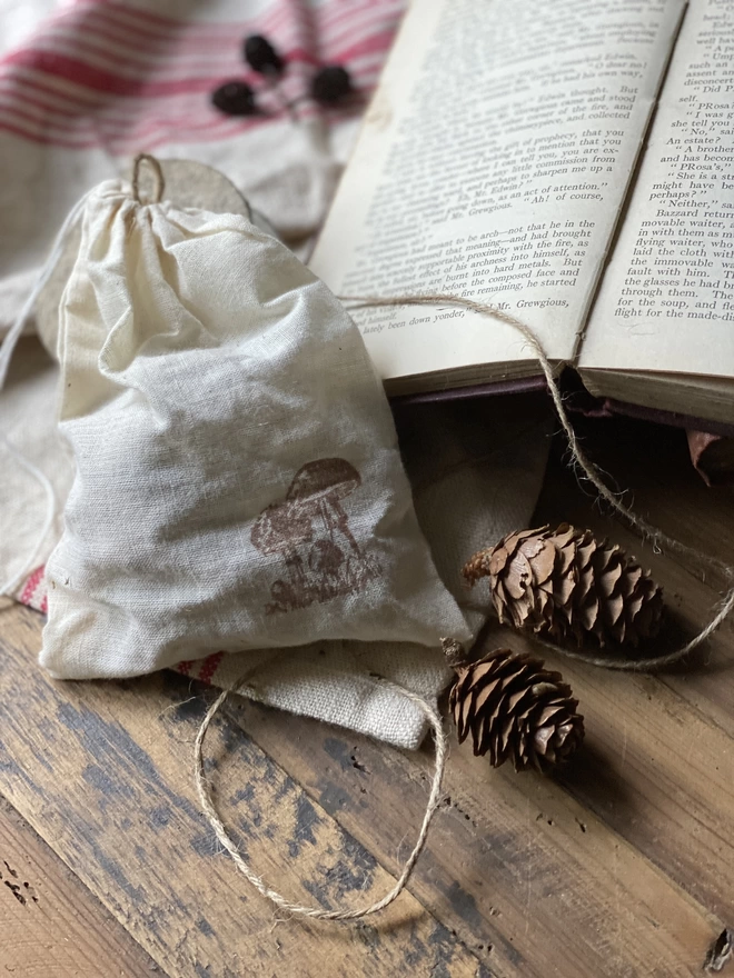 A white pouch with a small toadstool print in brown, on display next to an open book and some decorative pine cones.
