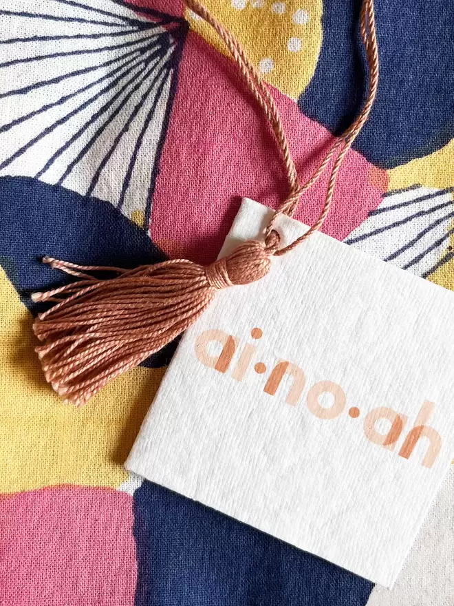 Detail of white recycled tag with ainoah brand logo and dusty pink tassel and hanging loop