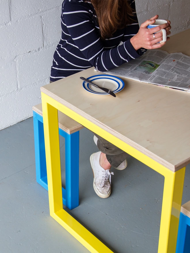 woman sat drinking coffee at a minimalist yellow-legged dining table and blue bench