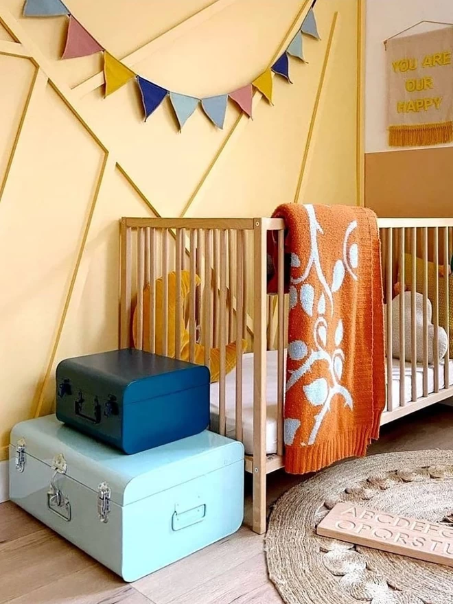 A wooden cot pictured in the corner of a warm yellow panelled nursery. The rust briar rose junior blanket is folded over the side of the cot, and 2 blue metal trunks are stacked at the end. Bunting in complimentary colours hangs on the wall above.