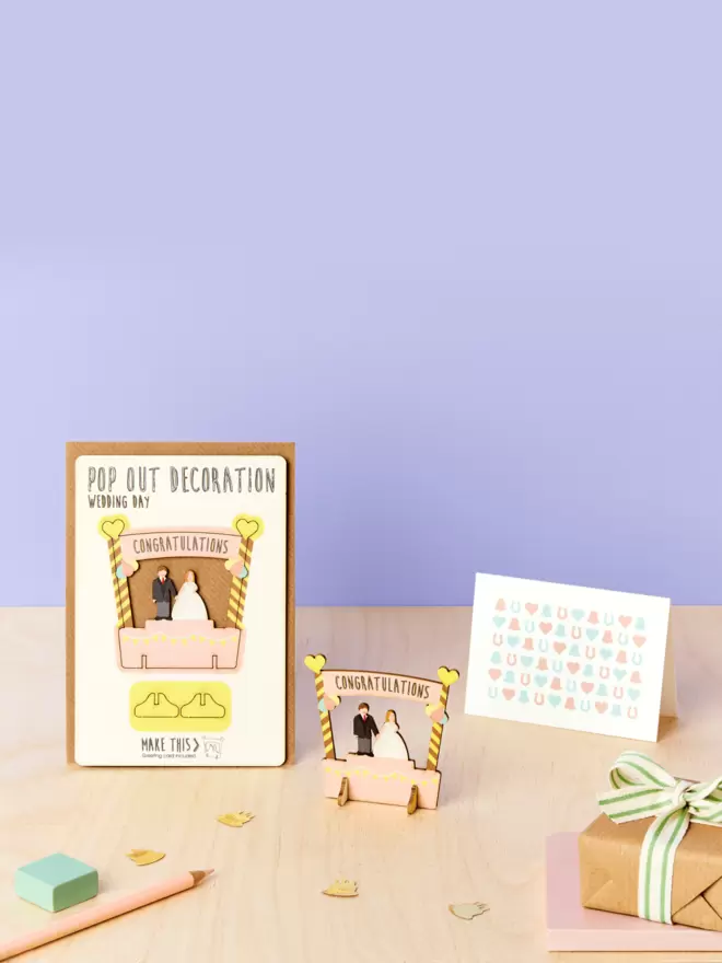 Wedding day congratulations 3D decoration and confetti pattern wedding card and brown kraft envelope on top of a wooden desk against a lilac coloured background