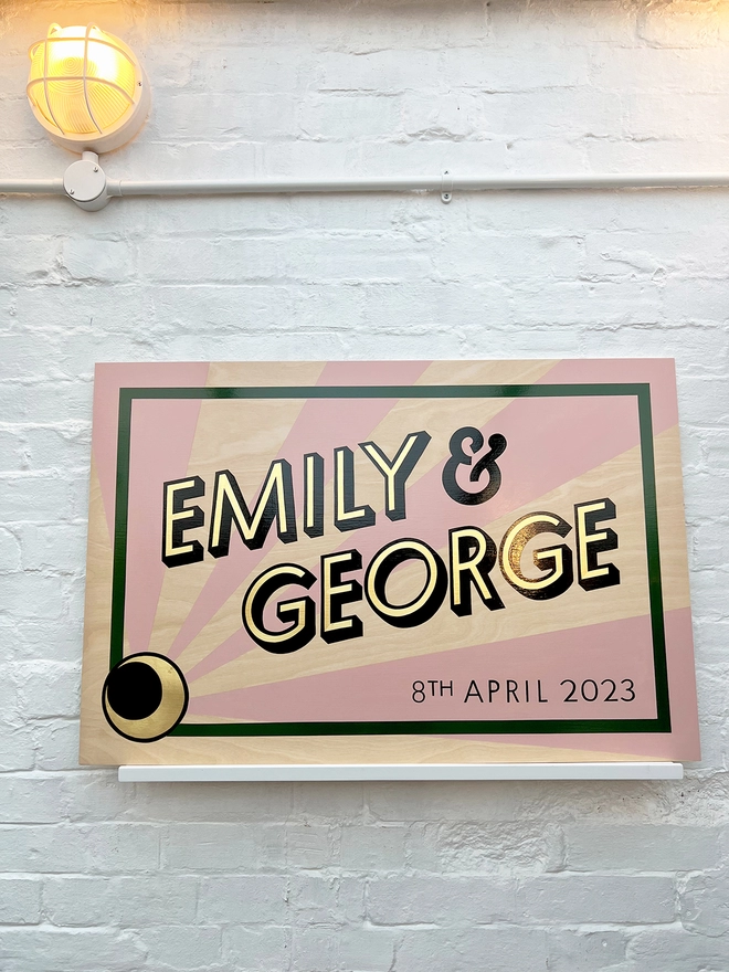 Modern wedding sign featuring the couple's names in gold leaf, outlined in black, and the wedding date, with a sunburst background and a border. Shown against a white brick wall, straight on. 