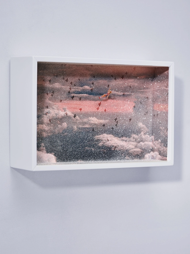 Miniature scene in an artbox showing a tiny woman flying with birds against a cloudy pink sunset sky 