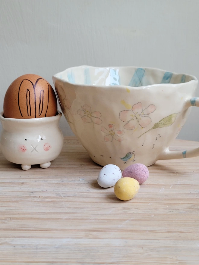 a handmade cup with blossom and bird painted on with a bunny rabbit egg cup with a brown egg and bunny ears next to it