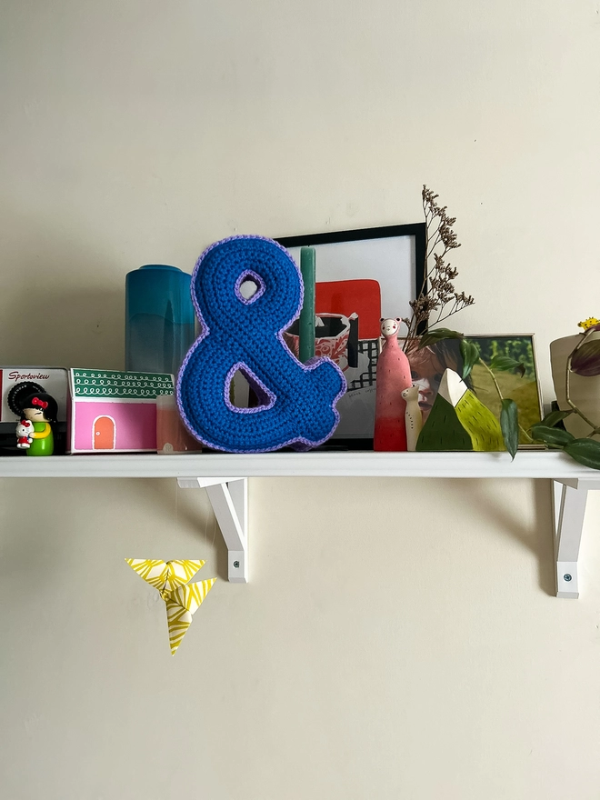 Crochet Cushion shaped like an Ampersand in Blue and Lilac sits on a shelf