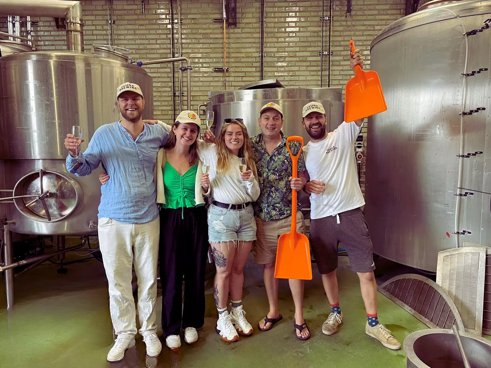 5 people celebrating in front of brewery tanks, wearing branded merchandise and holding brewing tools