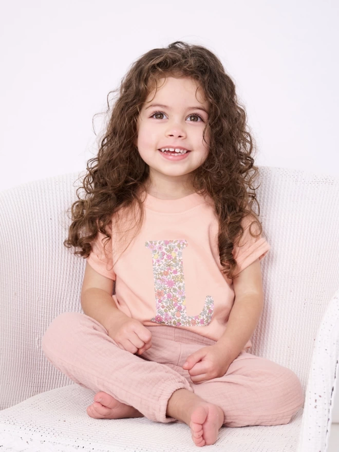 A smiling 3 year old girl in a pink t-shirt with her initial appliquéd in floral Liberty print 