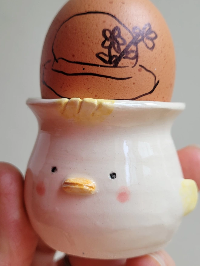 chick egg cup in hand made ceramic held in a hand with a brown egg inside and an Easter bonnet drawn on it 