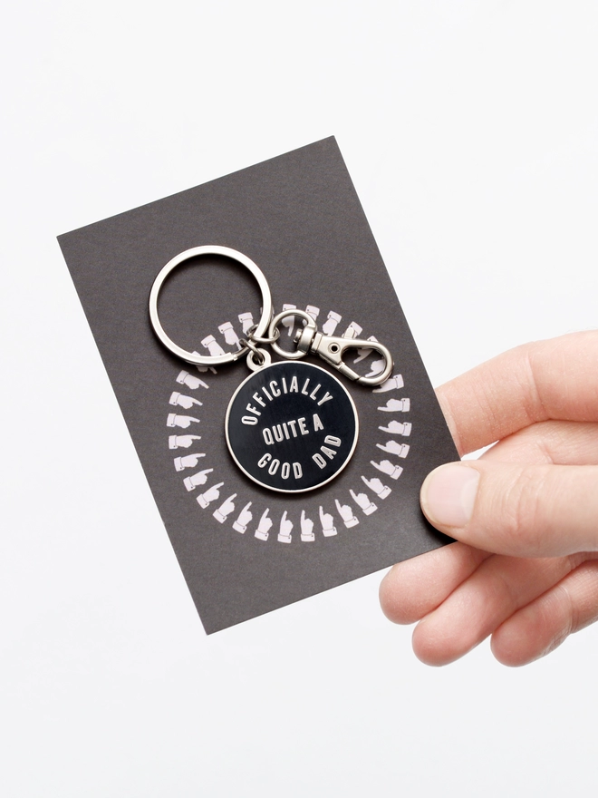 Hand holding out a Black and silver enamel keyring with 'Officially Quite a Good Dad' design