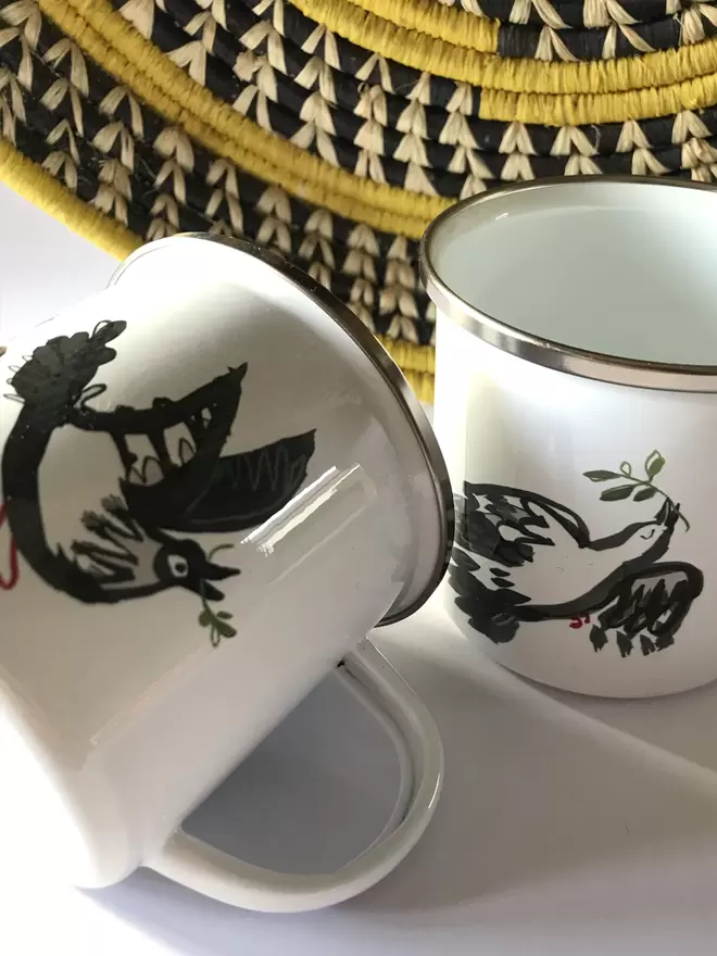 Two white enamel mugs with a black illustrated peace dove design, lying next to a black and yellow basket