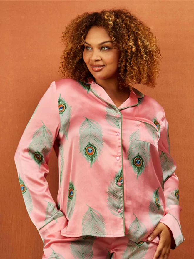 Model looks to left with her hand in the pocket of pink based satin pyjamas in a traditional classic shape with green printed peacock feather