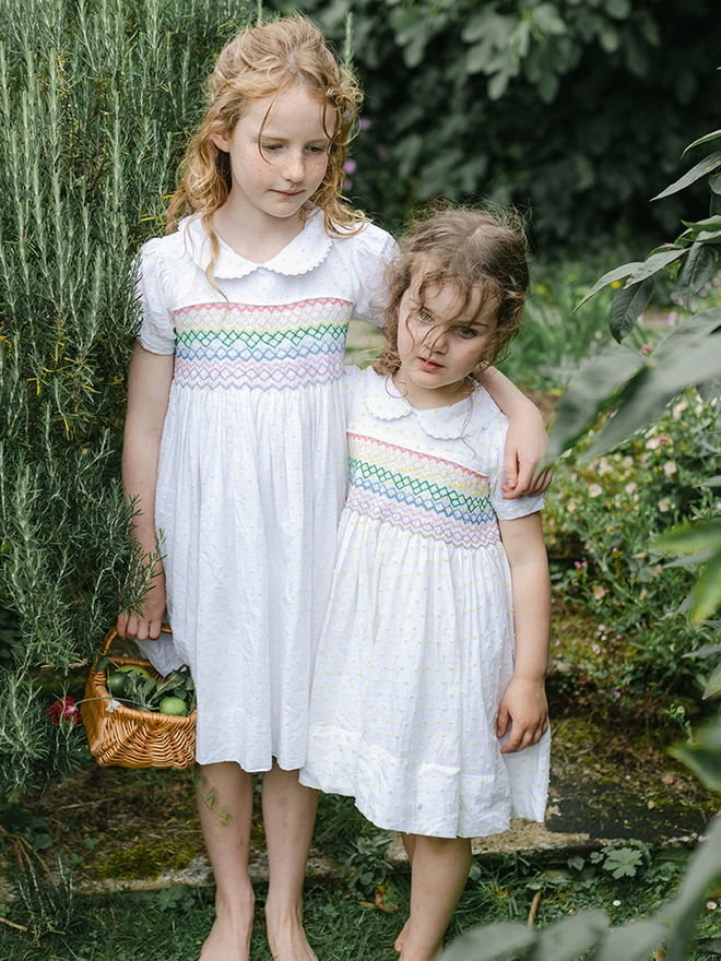 Two girls in white dresses with peter pan collars and rainbow smocking stand in a garden one with her arm around the other