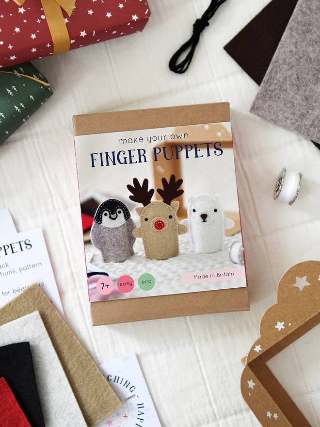 A craft kit in a brown cardboard box with a sleeve that features three winter finger puppets lays on a white surface with craft kit materials beside it.