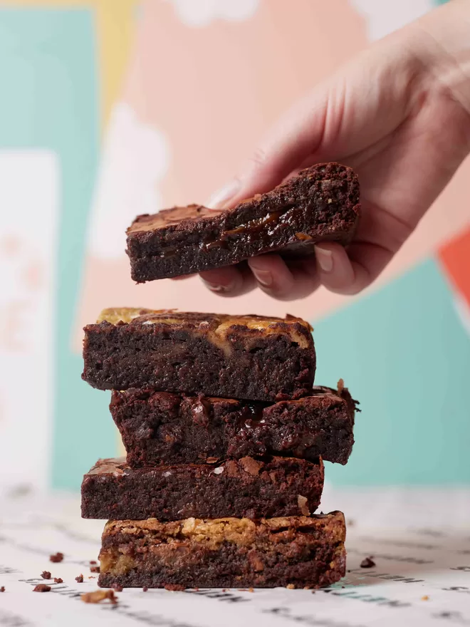 Mixed stack of Hetty's brownie originals with a hand adding another brownie to the top of the pile for scale against a colourful background