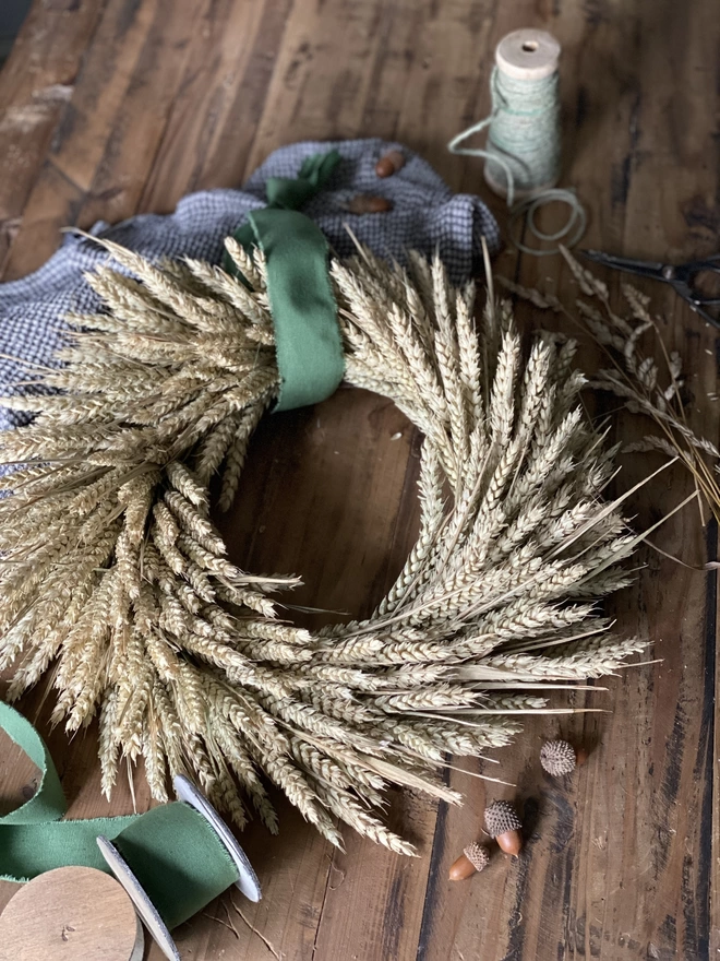 A handmade September Harvest Wheat Wreath with a sage green ribbon looped around the top, on display with a blue chequered cloth and small autumnal accessories