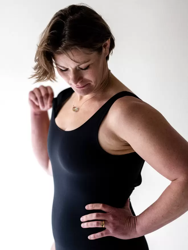 Woman with short brunette bob with hand on hip in studio looking downwards, wearing a Davy J Sustainable Waterwear black classic cross back swimsuit