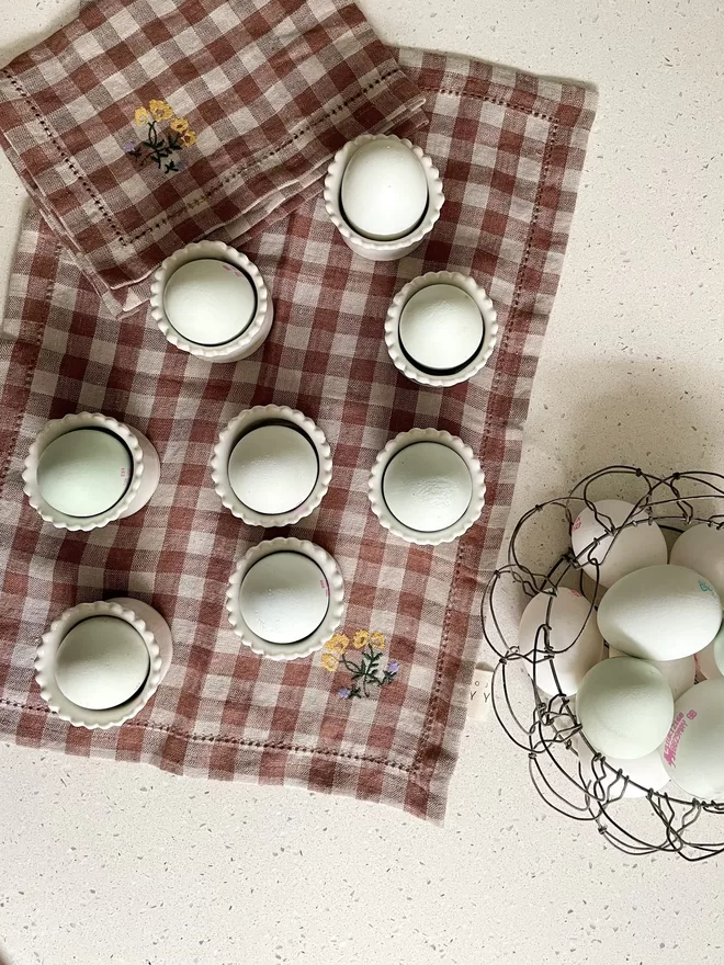 top view of scalloped edge egg cups with eggs in and a wire basket of eggs on brown checked placemat