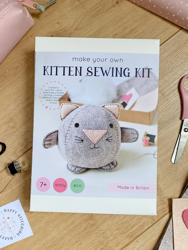 A craft kit box to make a felt kitten kit with the words "Make your own kitten sewing kit" lays on a wooden desk beside various felt and craft kit components.