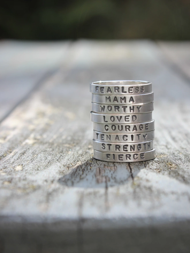 A collection of sterling silver simple plain band rings with words of empowerment stamped on them, stacked together on a weathered blue painted table.
