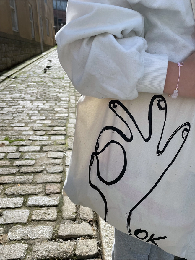  Black ink hand drawn in an OK gesture, on a white tote fairtrade bag. On the arm of a person in a cobbled street, afternoon light.