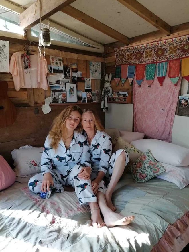 Mother and daughter sit up on a bed together surrounded by trinkets and soft furnishings wearing a fresh white based traditional pyjama with a watercolour hummingbird print