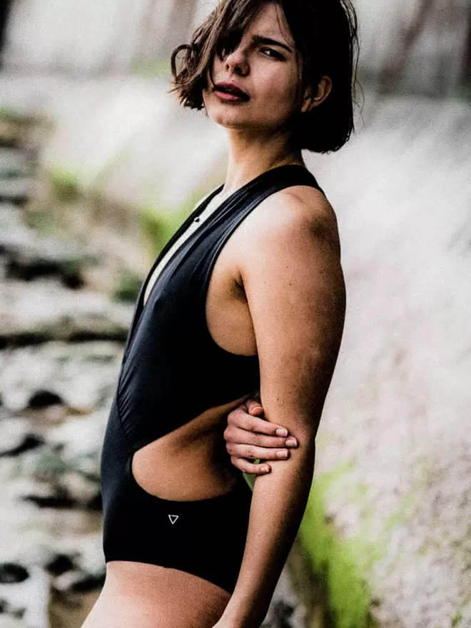 Woman facing side on, by sea wall with hair across her face and one arm behind her, wearing black Davy J Sustainable Waterwear cutout swimsuit with plunging neckline