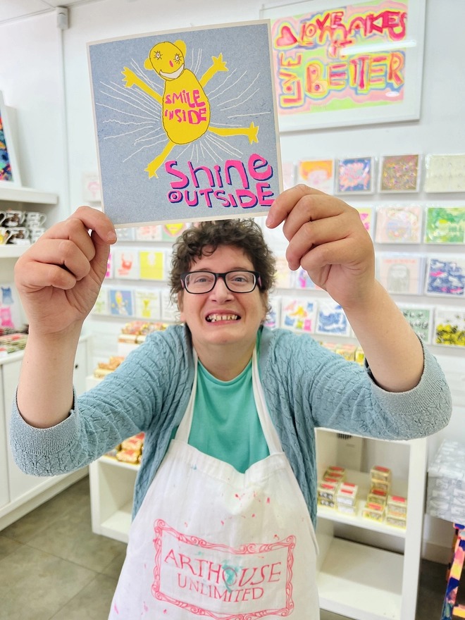 Artist holding the Smile Inside Shine Outisde card printed in blue, pink and yellow