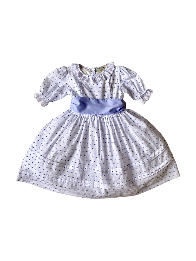A white dress with blue swiss dots with lace edged sleeves and neckline and a blue sash