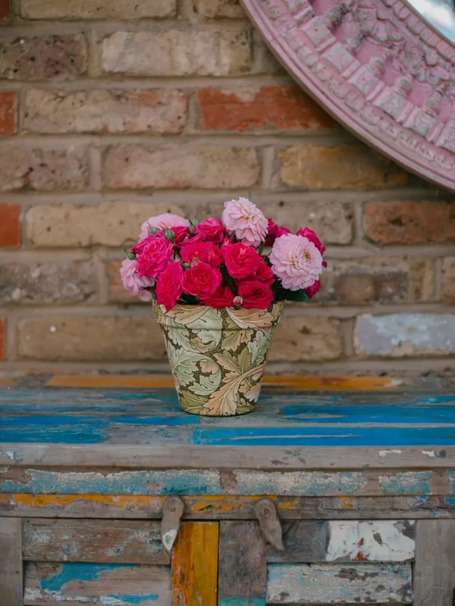 William Morris Acanthus Design Plant Pot seen with pink flowers on a blue wooden chest.