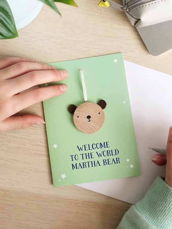 A green greetings card with a small wooden bear keepsake and the words "Welcome to the world Martha bear" printed on is on a wooden desk. 