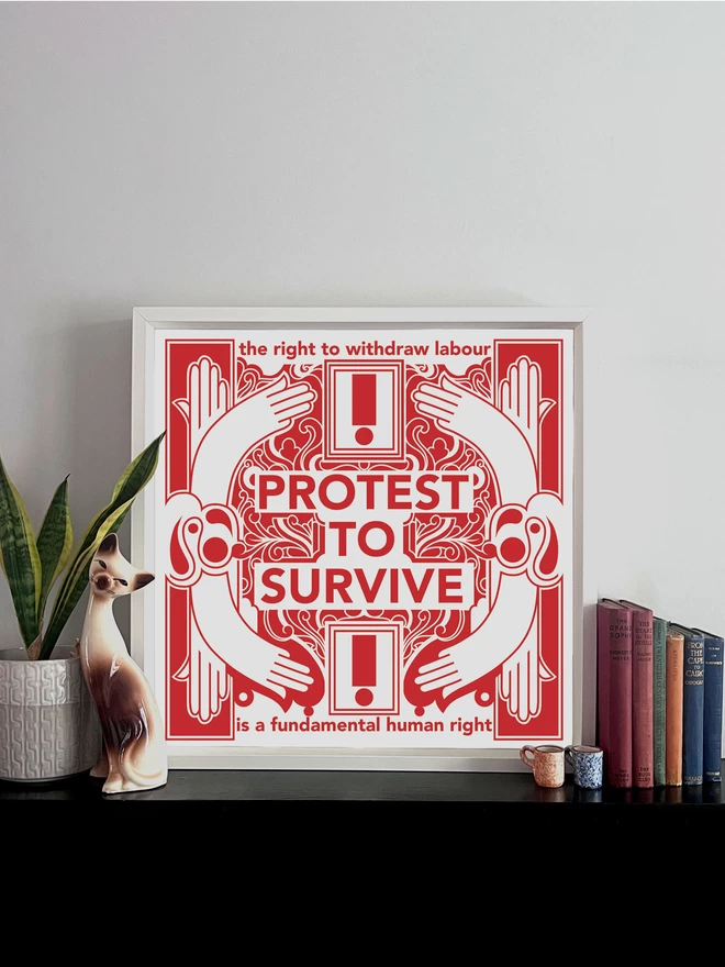 The bold red and white print with "Protest to survive" written at the centre, as well as “the right to withdraw labour” at the top, and “is a fundamental human right” at the bottom sits in a white frame, resting on a shelf with a pot plant, a cat ornament, and a collection of books