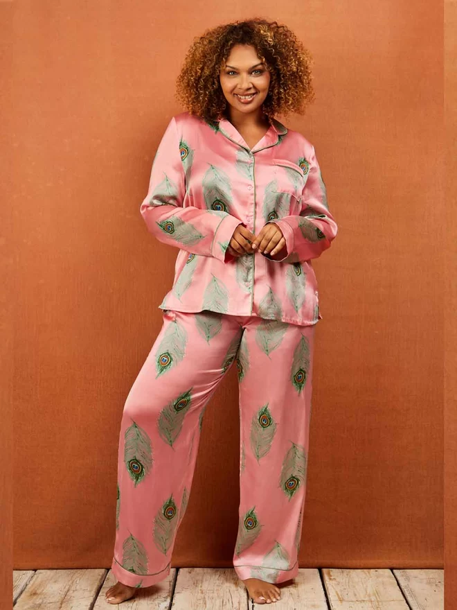 Model looks direct into camera wearing pink based satin pyjamas in a traditional classic shape with green printed peacock feather