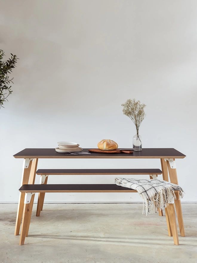side view of a stylish and minimalist dining set comprising a table and benches with black Fenix top, black coloured steel brackets and solid oak legs, laid for a simple lunch with a loaf of fresh bread
