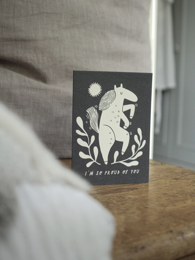 Black and white greeting card with illustration and the words I'm so proud of you written on it stood up on wooden surface with grey cushion behind it 