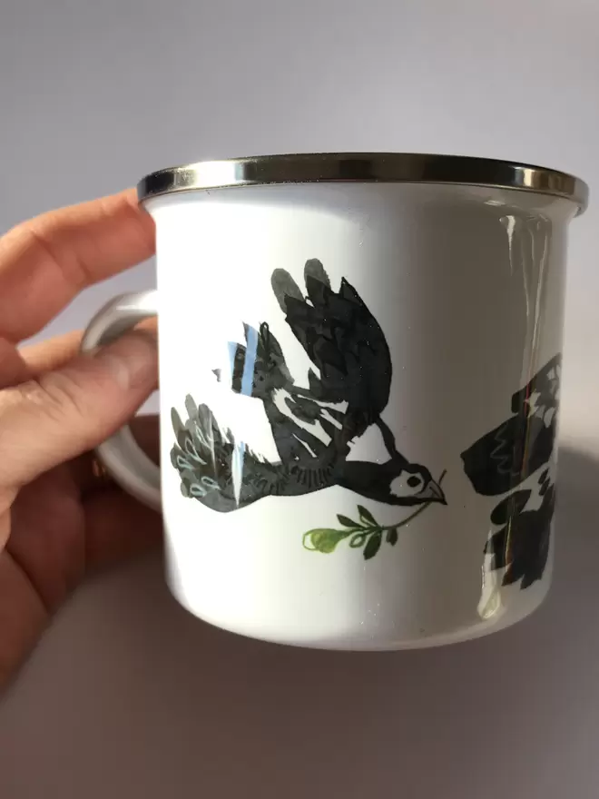 A hand holds a shiny white enamel mug decorated with an illustrated back dove design