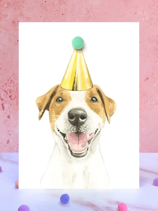 A greeting card featuring a hand painted design of a jack russell terrier, stood upright on a marble surface surrounded by pompoms. 