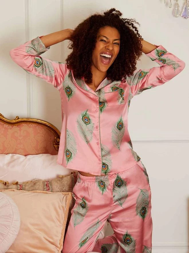 Model stands, arms in her hair, grinning, wearing pink based satin pyjamas in a traditional classic shape with green printed peacock feather