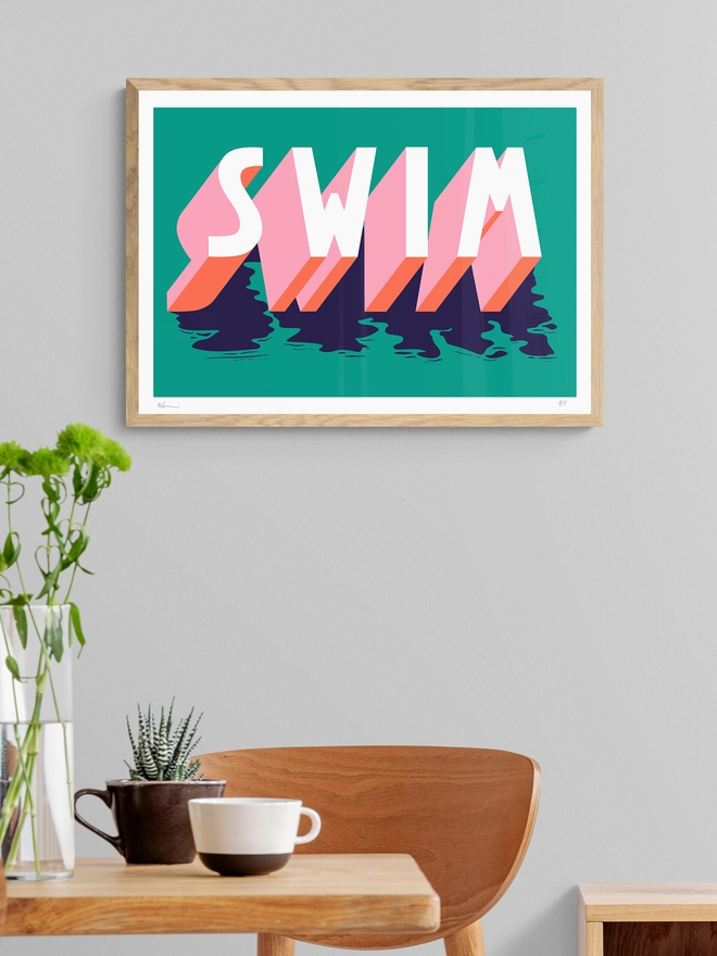 Framed screenprint of the word SWIM in 3d typography in pink, orange and blue on a green background by artist Survival Techniques. The frame hangs in a dining room. 