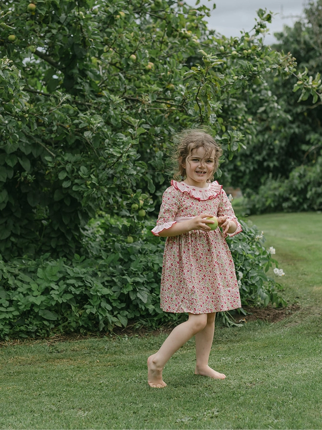 A little girl is surrounded by flowers wearing a floral dress with a pink frill collar and hand embroidered detailing