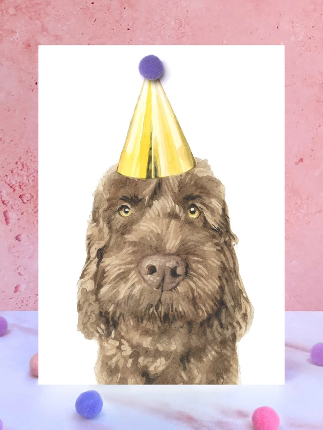 A greeting card featuring a hand painted design of a chocolate cockapoo, stood upright on a marble surface surrounded by pompoms. 