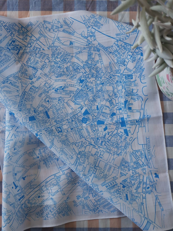 A lifestyle photo of a Mr.PS Sheffield Map hankie printed in sky blue with a plant pot on a gingham tablecloth.