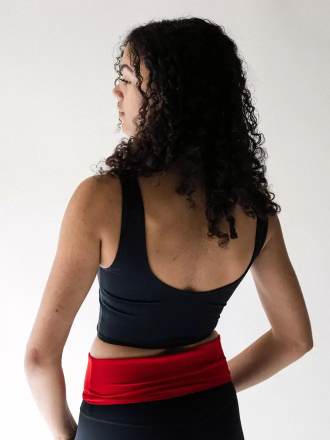 The back view of a lady with long black curly hair wearing a Davy J Sustainable Swimwear black cropped swim top with a scoop back and shoulder straps and a pair of black high waist briefs with the waist folded down, showing the red lining